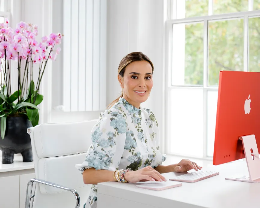 Smiling woman in floral blouse sitting at white desk using a red iMac computer with a potted orchid in the background.