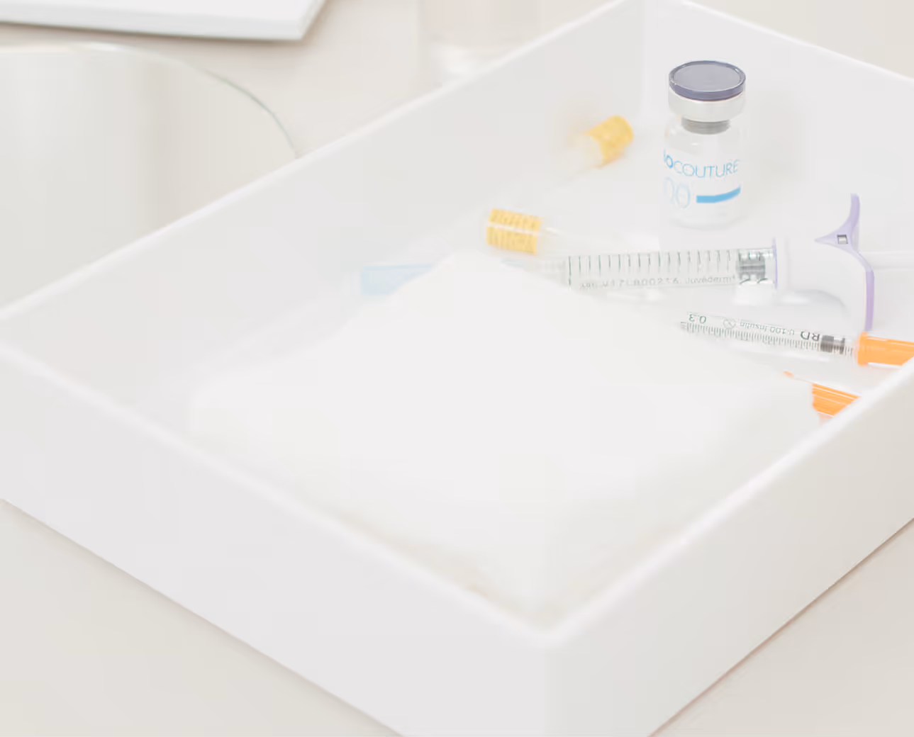 White tray containing syringes, a small vaccine vial, and capped needles on a medical surface.