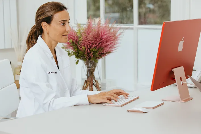 Female doctor in white coat typing on keyboard at modern desk with orange Apple computer and flower vase in background.