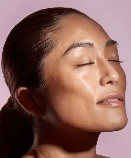 Close-up of a woman with smooth glowing skin and closed eyes against a soft pink background.