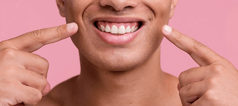 Close-up of a smiling person pointing at their teeth against a pink background.