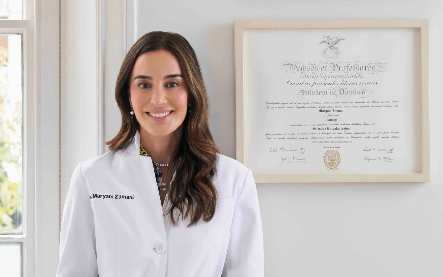 A smiling woman wearing a white medical coat with the name Maryam Zamani embroidered, standing next to a framed diploma on the wall.